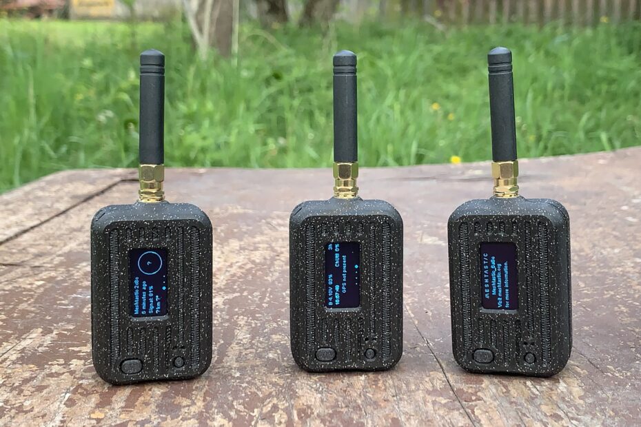 Trzy małe radia Meshtastic, czarne, z niewielkimi antenami podstawowymi. Na strym drewnianym stole w ogrodzie.